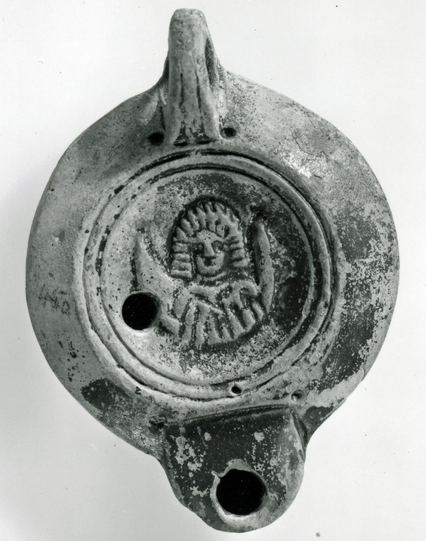 Mould-made pottery lamp with a circular body, a rounded shoulder, and a short flat-topped semicircular nozzle set into the shoulder and defined by a lateral groove. At the rear is a pierced handle, grooved along the upper edge. The discus is surrounded by two circular grooves, the outer one pierced with an air-hole. On the discus is a draped bust of Luna with the horns of a crescent moon rising on each side of her. Across the slightly raised base is the impressed name CATILIVEST. Covered with a…