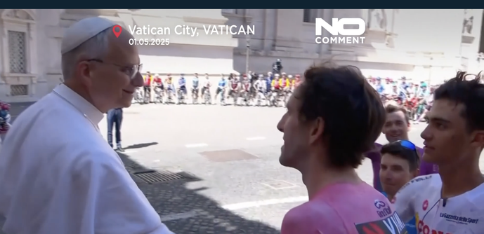 The image depicts a scene in Vatican City on May 1, 2025. In the foreground, a man dressed in white religious attire, likely a high-ranking clergy member, is seen interacting with a group of cyclists. The man is facing the cyclists, who are wearing colorful cycling jerseys, with one cyclist prominently in a pink jersey, a common color for the leader in the Giro d'Italia. The background shows a line of cyclists, suggesting a cycling event, possibly the Giro d'Italia, passing by a historic buildi…
