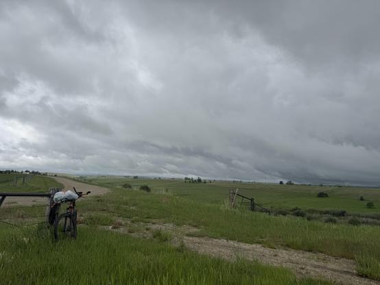 A pink mountain bike is leaning against a fence post next to a gravel road under low fast moving storm clouds.