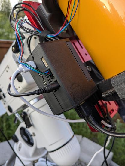 A black box with wires plugged into it. In the background is the yellow telescope from the previous pictures. The multicolor wires enter the frame from the top left and plug into the black box. Below the telescope, a telescope mount can be seen, and behind that a yard that needs to be weeded.