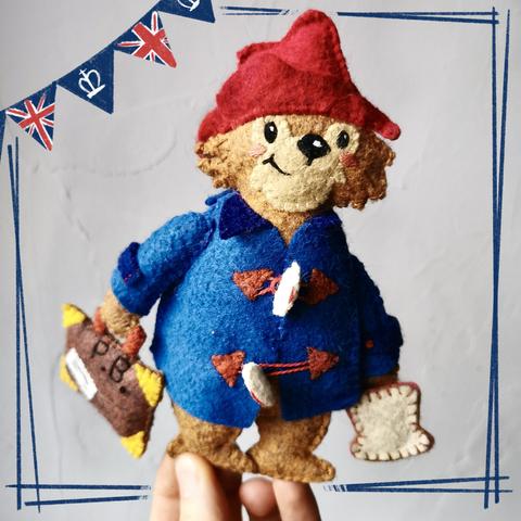 Picture of a little handmade Paddington Bear plushie held in front of a white wall. 
Made of felt, he is wearing his signature blue trench coat and red hat and holding his suitcase as well as a marmelade sandwich. 