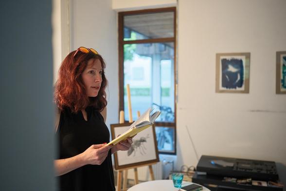 Nahaufnahme von Fee, sie blickt ins Publikum und hält ihr Buch. Im Haar, wie fast immer, eine Sonnenbrille. Im Hintergrund eine Collage von ihr auf einer Staffelei.