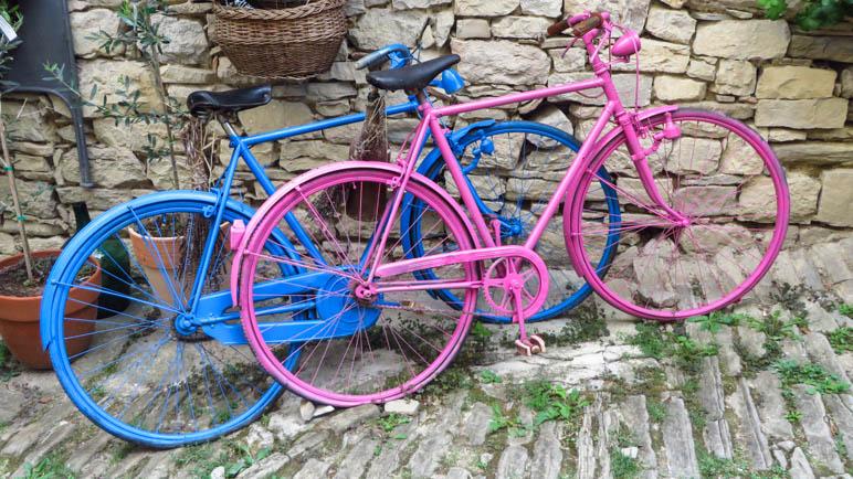 Zwei ältere Herrenfahrräder, die aneinander gelehnt in einem sehr alten Ort stehen. Das hintere Fahrrad ist, bis auf den Sattel, komplett blau eingefärbt. Das vordere, ebenfalls abgesehen vom Sattel, komplett rosa.