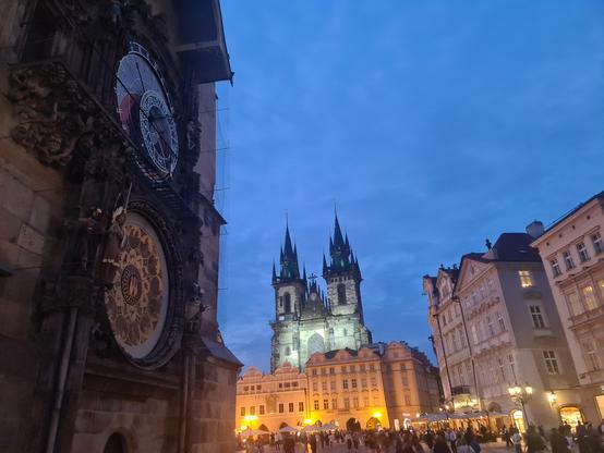 A photo of Prague in the evening. In the foreground is the old townhall, the location of the welcome reception. The antique clock of the townhall can be seen. There are crowds of people and further buildings, including a church with two towers, in the background.