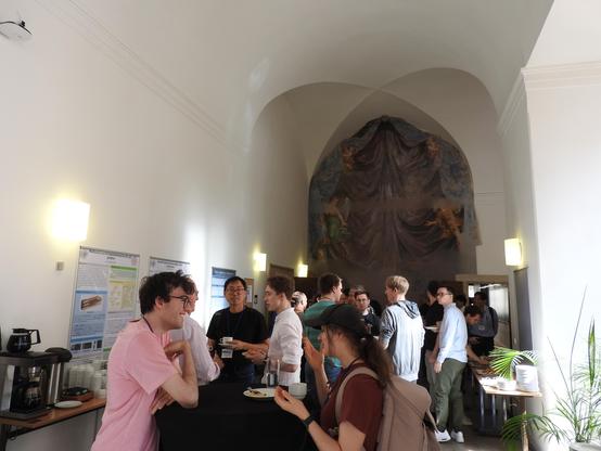 A photo of a coffee break at <Programming> 2025. The room has a high ceiling and the wall in the back has a big painting on it. In the foreground, people are discussing and eating snacks.