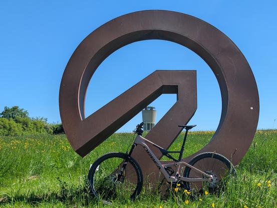 Mountainbike vor einer Skulptur auf einer Wiese im Schönbuch. Im Hintergrund ein Wasserturm.