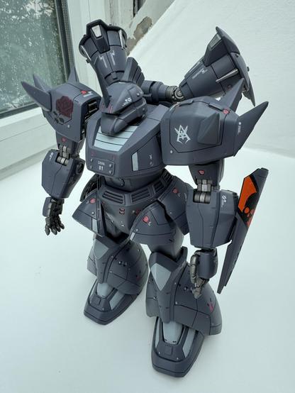 A detailed model of a mecha robot stands on a white surface, featuring a dark gray color scheme with decorative elements like a rose and insignias. The robot is equipped with various mechanical parts, including shoulder pads and a shield, showcasing intricate craftsmanship.