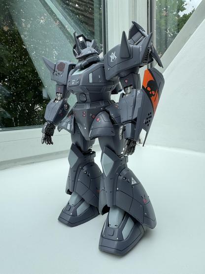 A detailed model of a robotic figure stands on a white surface near a window. The figure features a predominantly gray color scheme with spikes on its shoulders, intricate mechanical details, and decorative elements like a rose and a skull on its shield. 
