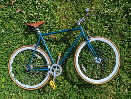 A metallic blue green bike with a cognac leather saddle and yellow pedals laying in a clover field.