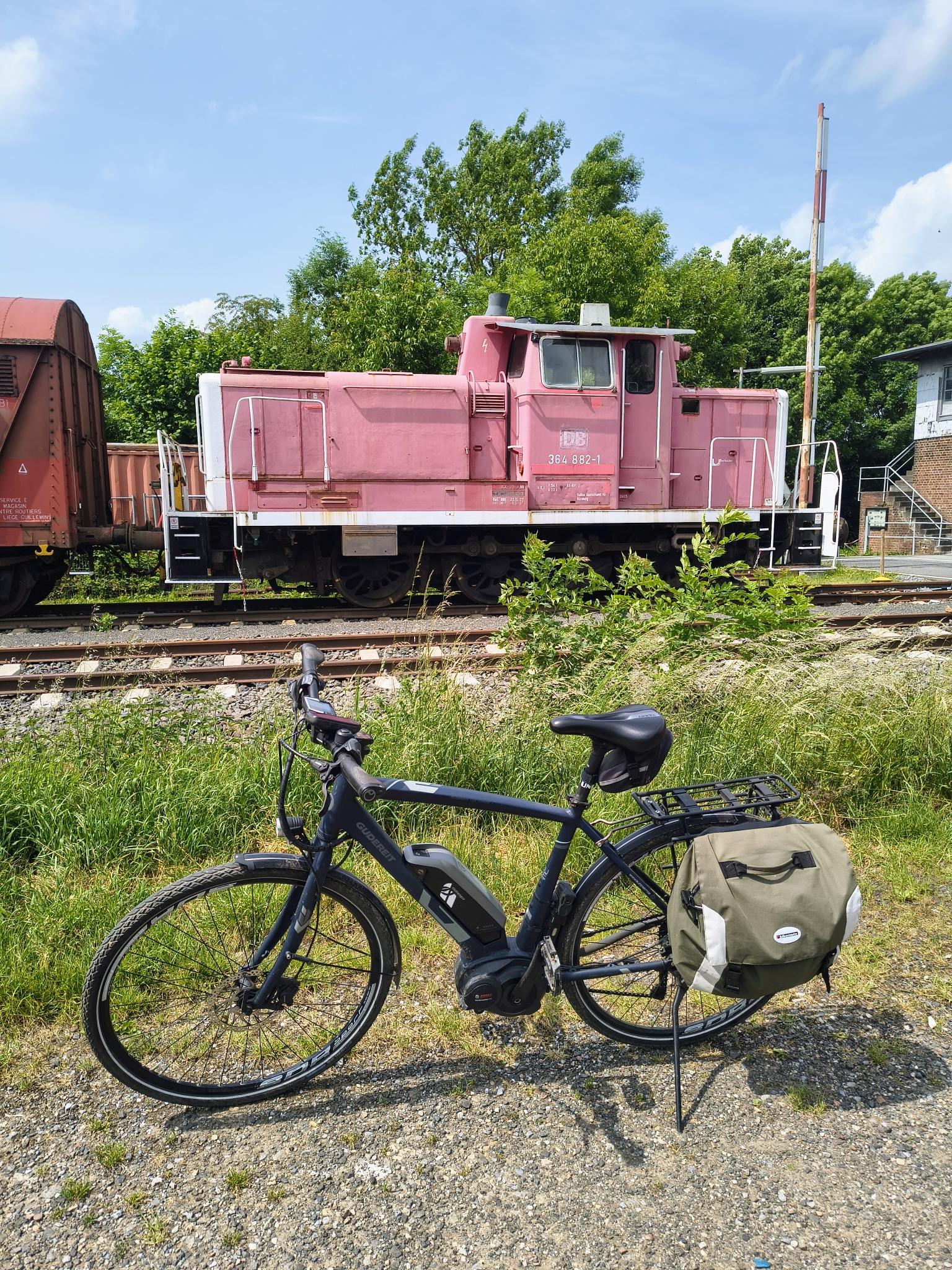 Ein Trekkingrad (eBike) steht vor Schienen. Im Hintergrund steht eine alte Rangierlok, deren rote Farbe sehr ausgeblichen ist.