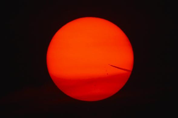 Photograph of a bright red and orange Sun with its light filtered through smoke and ash from the Canadian wildfires. A jet and contrail are visible headed towards the center from the right side. Sunspots are visible below the jet.