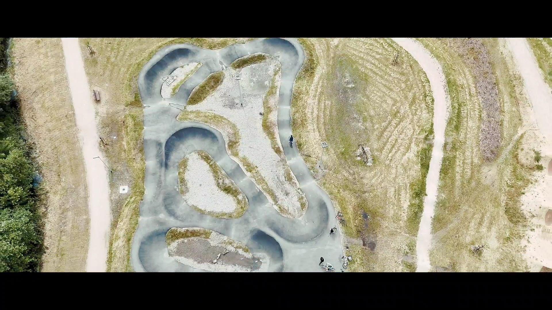 Bildbeschreibung von KI:
Das Bild zeigt von oben eine geschwungene, asphaltierte Strecke, die in viele Kurven, Hügel und Wellen unterteilt ist. Die Bahn ist grau und hebt sich deutlich vom umgebenden, blassgrünen und braunen Grasland ab. Dazwischen sind helle, mit Kies gefüllte Flächen sichtbar, die wie Inseln innerhalb der Strecke wirken. Die Form der Bahn ist unregelmäßig und verläuft wie ein verschlungener Pfad mit vielen Schleifen.
Rund um die Strecke führen breite, helle Kieswege durch eine offene, parkähnliche Wiesenlandschaft. Am unteren Bildrand (Süden) stehen ein paar Menschen mit Fahrrädern oder Rollern – vermutlich Kinder oder Jugendliche. Im linken Bildbereich sind auch einige Bäume zu erkennen, die einen angrenzenden Wald andeuten.
Insgesamt vermittelt das Bild eine aktive, sportliche Umgebung an der frischen Luft, vermutlich in einem Freizeitpark oder an einem Spielplatz.

