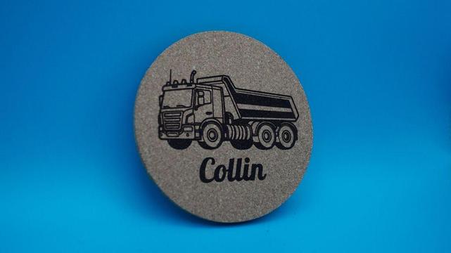 Korkuntersetzer mit graviertem Kipplaster und dem Namen „Collin“, vor blauem Hintergrund schräg fotografiert.