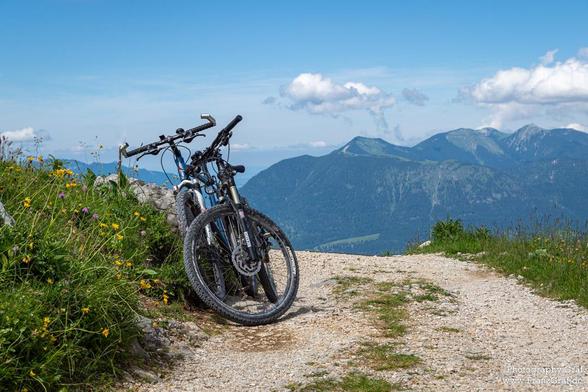 Two bicycles are parked on a dirt road in a scenic outdoor setting. The dominant colors in the image are shades of green, with a clear blue sky in the background. The bicycles are in good condition, with one having its wheel prominently displayed in the foreground. The image shows a cycle and wheel as identified objects. The scene suggests a peaceful mountain biking excursion, with the bicycles ready for use. The setting is serene, with no people visible in the frame. The overall composition ev…