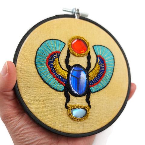 Broderie sur une toile orange clair tendue sur un cercle à broder en bois noir.
Elle représente un scarabée ailé tenant un soleil et une demi-lune entre ses pattes, incarnation du dieu antique égyptien Khépri.
Le soleil, le corps du scarabée et la demi-lune sont des morceaux de CD fixés à la toile avec la technique de la broderie miroir, puis recouverts de résine époxy teintée.
Beaucoup de fil doré a été utilisé.