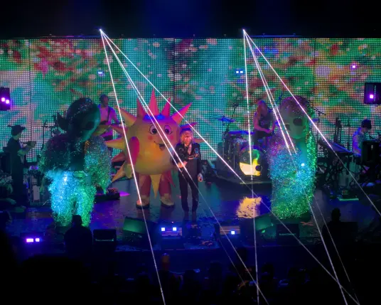 Photography of The Flaming Lips playing onstage. Besides the band, two people in tall alien costumes and one in a sun costume are onstage. 
