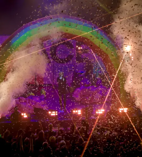 Photography of The Flaming Lips playing onstage. Wayne Coyne is singing under a confetti rain and an inflatable rainbow, surrounded by vertical smoke cannons on his left and right.