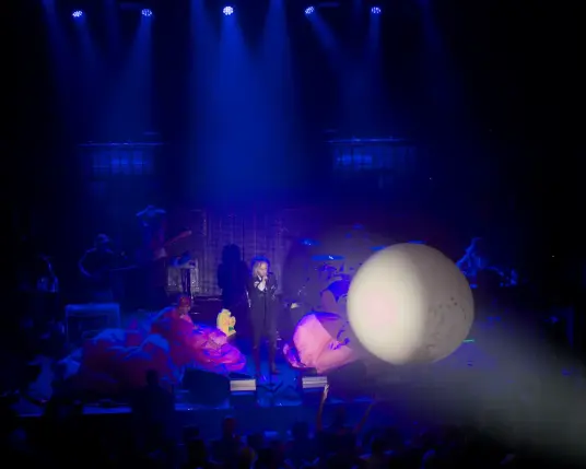 The Flaming Lips onstage. Wayne Coyne is speaking under blue lights while a large white balloon is floating in front of him over the public, under white light from a projector.