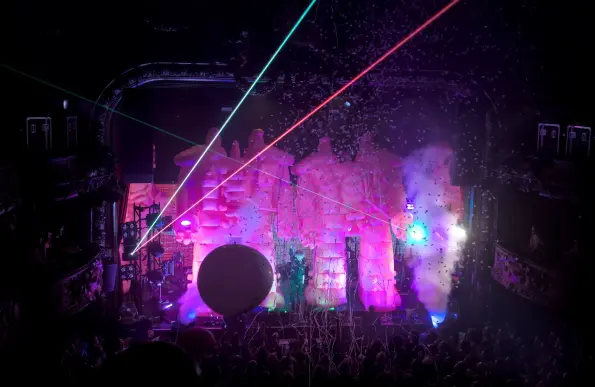 The Flaming Lips performing onstage. Wayne Coyne is singing between two giant pink inflatable robots under a confetti rain, with a vertical smoke jet on the right side. A large balloon is bouncing over the public.