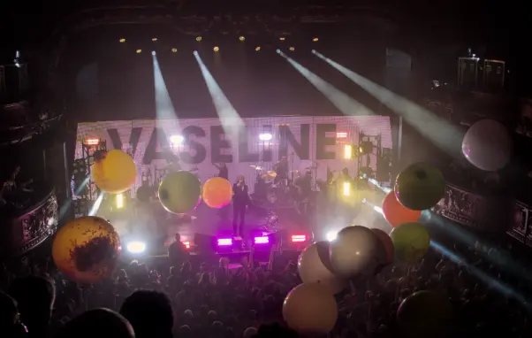 The Flaming Lips performing onstage. The back screen displays the word "vaseline" while many large balloons are bouncing over the public in the classical-style concert room.