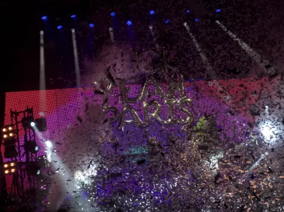 The Flaming Lips performing onstage. A balloon shaped like letters forming "Fuck Yeah Paris" is brandished by Wayne Coyne, hidden behind a thick confetti rain.