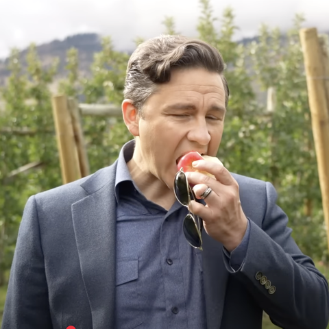 A dweeb of a man is standing outdoors, wearing a dark blue blazer over a matching blue shirt. He is holding a red apple in his right hand, taking a bite. His left hand is holding a pair of sunglasses. The background shows a blurred natural setting with greenery and wooden posts, suggesting an orchard or a similar outdoor environment. The man has short, graying hair and is wearing a ring on his left hand. The lighting is natural, indicating it is daytime. The Sod - Pierre Poilievre-  lost the Ca…