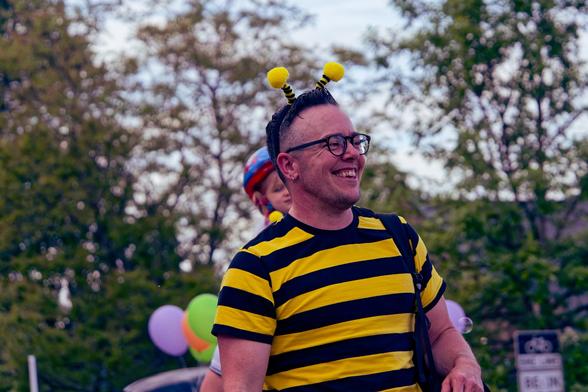 Man wearing a yellow and black striped short sleeve shirt with antenna headband and thick glasses.  Behind him are some balloons and a child wearing a bicycle helmet