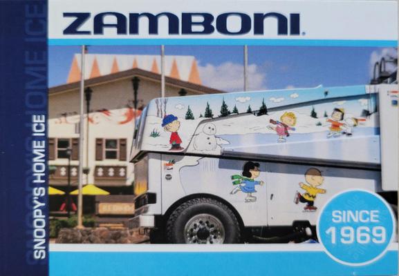 Promotional card showing a daytime exterior photo of a Zamboni ice-resurfacing machine in the plaza in front of the Snoopy's Home Ice rink in Santa Rosa, California. The Zamboni is illustrated with cartoon scene of characters from Peanuts, as they skate on an outdoor winter pond. The Zamboni wordmark appears at top in rounded navy block letters, on a white horizontal background row. A navy vertical border in the left has text Snoopy Home Ice in white block letters. A circular light-blue bubble at lower-right has text Since 1969 in white block letters. The card has a light-blue bottom border.
