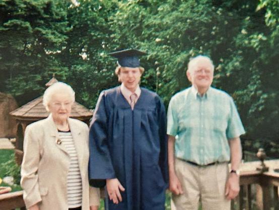 I'm standing in a graduation robe and cap with my grandmother on the left in a white suit and my grandfather on the right in a teal shirt and khaki pants. My mouth is contorted as I was talking to them at the time the picture was taken and it's the only one from graduation with my grandparents in it! Sunflowers blossom in planters along the railing and a gazebo is in the background.