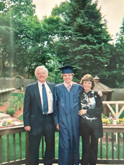 I'm standing in a graduation robe and cap with my father on the left in a suit and tie and my mom on the right in a black blouse with a white design on the front. We are standing on a deck with our backyard behind us in full spring bloom as well as an above ground pool in the back left.