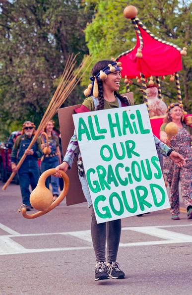 A woman wearing a headdress with little gourds attached and holding one in her hand is wearing a sign that says ALL HAIL OUR GRACIOUS GOURD in green letters on white card before her,  behind and to the left is a man with similar headware carrying a large bundle of sticks or straw and behind and to the right can be seen a huge gourd in a red palanquin
