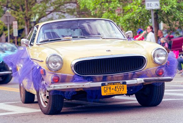 A class yellow Volvo in light yellow with round headlights and a rounded-off grille has the person obscured by reflections but i can swar I can see the ears of a dog in the passenger seat,  it's wrapped up in a yellow tutu and has new york plates with a few spectators and a sign that seems to read BE IN in the background