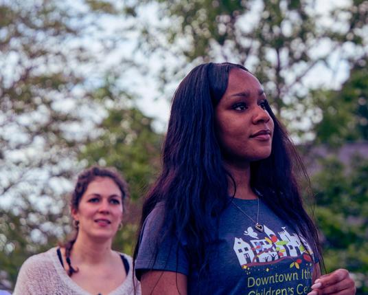 In the foreground a black woman with long dark hair and a little heart necklace wears a dark grey shirt for the Downtown Ithaca Children's Center while in the background a white woman with brown hair is looking the same direction wearing a white blouse with a large hole for the neck and behind that find out of focus trees