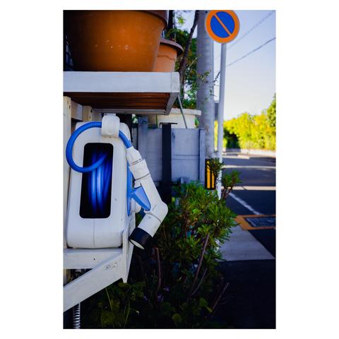 A close-up of a white and blue hose reel mounted on a low outdoor shelf in a residential area of Nagoya. The vibrant blue hose stands out against the soft shadows of early morning or late afternoon. Terra-cotta planters sit on the shelf above, and a small bush partially frames the setup. In the background, a “no parking” sign leans slightly, and the quiet street stretches toward dense greenery.