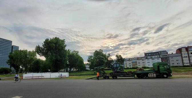 Asphaltfläche, dahinter ein mit Bauzäunen und weißen Planen abgesperrter Bereich. Von einem Sattelschlepper wird ein Bagger entladen.