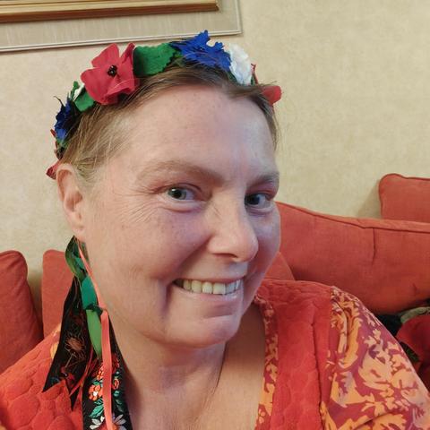 A white middle aged woman in an orange dress is wearing a wreath made from various textiles. It is covered in bright green birch type leaves, and fabric flowers in blue, red, and white. It has ribbons hanging from it in the back