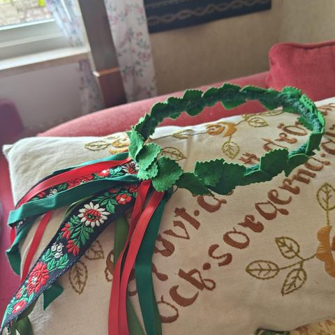 A wreath made from various textiles is lying on an embroidered cushion. It is covered in bright green birch type leaves, and has ribbons hanging from it. Red shiny ribbons, green cotton tape, and a jaquard ribbon with red and white flowers, and green leaves, on a black background.