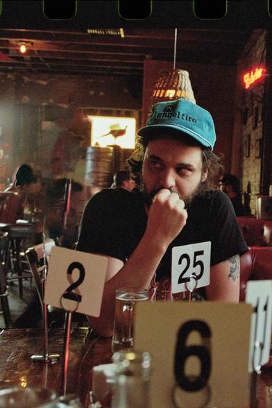 Max Knouse in blue cap sits pensively at a bar, surrounded by table numbers. Warm lighting and a casual atmosphere.