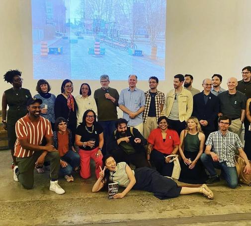 A group of diverse individuals poses for a photo, with some sitting and others standing. They gather in front of a large screen displaying an urban scene. Several people hold drinks, and one woman is lying on the ground playfully. 
