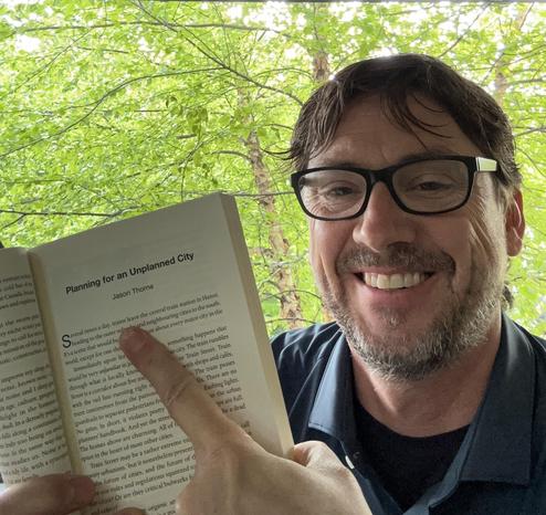 A picture of me holding an opened book turned to a page with a chapter titled "Planning for an Unplanned City" by Jason Thorne.