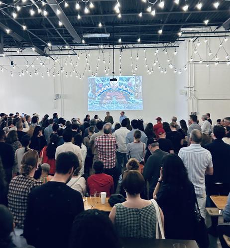 A large crowd is gathered in a well-lit space, facing a projection on the wall displaying colorful artwork. The audience is comprised of people of diverse backgrounds, engaged with the presentation. Decorative string lights hang from the ceiling.