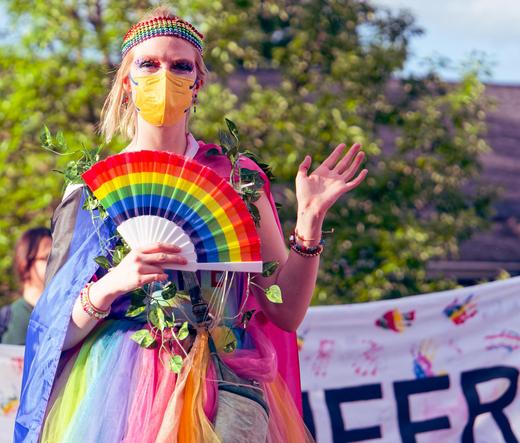 Person wearing rainbow colored clothes and a rainbow headband and holding a rainbow fan with a yellow facemask behind a sign and spreading tree
