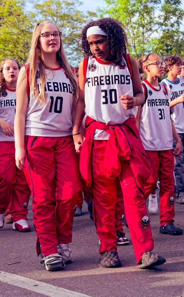 Basketball playing girls with red pants and white tank tops that say FIREBIRDS.  In fron two girls are prominent,  #10 who is white and blonde with long hair and glasses,  next to her #30 who is black with tightly curled hair and a white headband.