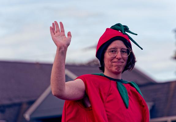 Woman wearing a big red tomato suit with green leaves around the neck has her hair mostly covered by a red tomato cap with a sprig of leaves,  holding one hand up.  Behind find a blurry building with a complex roofline and an sky with light clouds