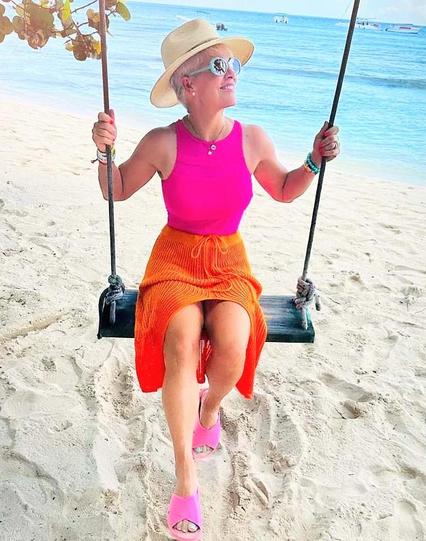 Older woman on a swing on a beach sand and water and she has bright beach clothes on hot pink shirt and sandals and an orange skirt and a straw hat and big white sunglasses 