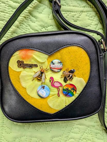 Photo of a black purse with a heart shape window on its one face. The window has a background of yellow felt and fabric flowers with several one-inch and enamel pins attached that include: the word "Catawampus" in a script font in a gold tone, two realistic jewel-style bees, a pink flamingo, two palm trees in a circle with a rainbow background, and two summer-style Anomaly pins.