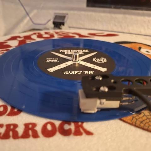 Eine blaue Schallplatte mit dem Titel "TOURIST TRAP" von "POOR IMPULSE CONTROL" wird auf einen Plattenspieler gelegt. Das Bild zeigt die Nadel und den Tonarm des Plattenspielers.