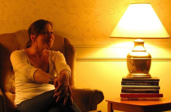 learn from personal stories: a photograph of a woman storyteller sitting in an armchair with a lamp on top of a pile of books on a table next to her