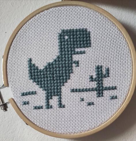 Zu sehen ist ein weißer Stickstoff, der in einem hellbraunen Holzrahmen eingespannt ist. In dunkelgrünem Garn ist jener Dino gestickt, der im Google Chrome Browser angezeigt wird, wenn man offline ist. Das gesamte Motiv ist mit einem Kreuzstich gestickt.