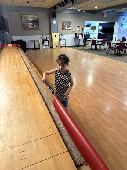A young child in a checkered shirt is interacting with a bowling-style lane in a large, empty room with wooden flooring. In the background, there are tables and chairs, along with wall decorations including paintings and a clock.